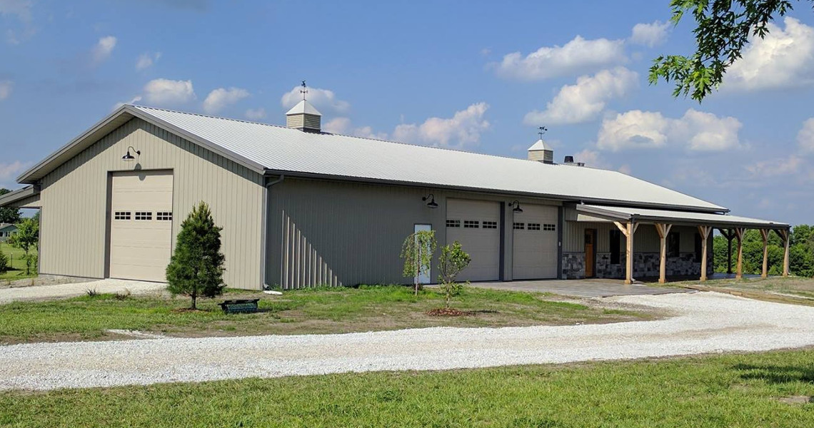 A large, beige metal building with three garage doors sits on a grassy field, embodying the charm of a Duncan dream home. A gravel driveway curves in front, while the sky is partly cloudy and a few small trees are scattered around the property.