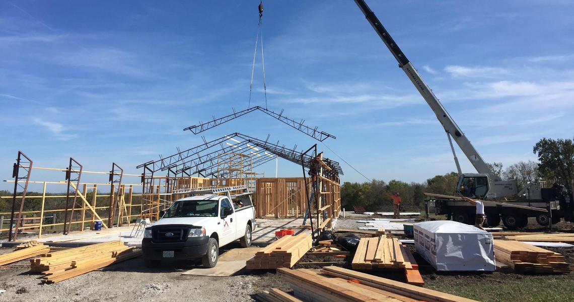 A construction site with a crane lifting a steel roof frame onto Duncan's dream home. A white truck is parked in the foreground, surrounded by stacks of lumber. Scaffolding is set up on the left, and the sky is clear and blue.