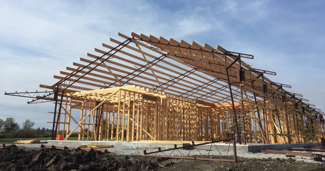 A wooden framework of a large building, perhaps someone's dream home, rises proudly on a concrete foundation. The structure, featuring beams and trusses against a blue sky in Duncan, is surrounded by dirt and construction materials, hinting at the bustling activity of its creation.