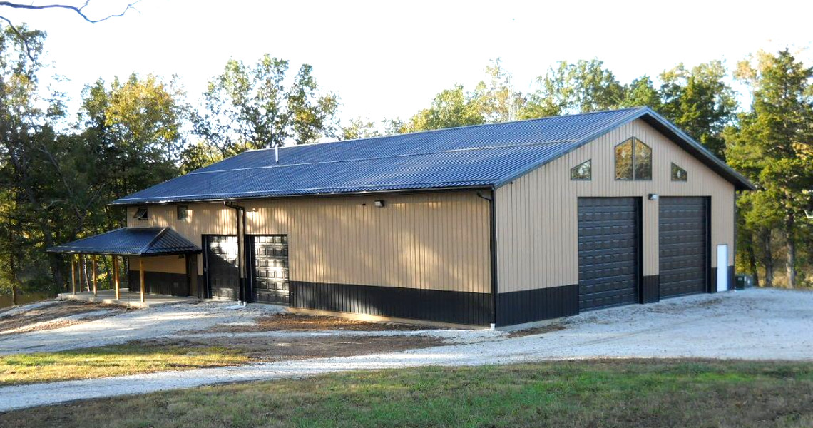 A beige and black metal building with two large overhead doors and a small covered entry area, this mountain getaway is nestled among trees. The structure, perfectly suited for an escape, sits on a gravel driveway against a clear blue sky backdrop.