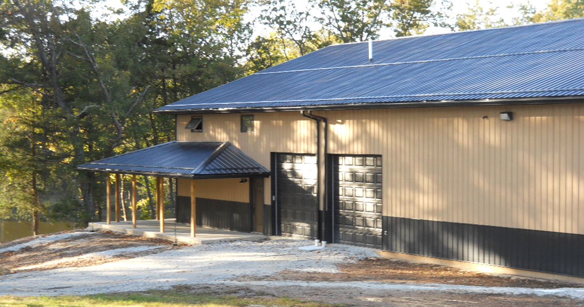 A beige and black metal building with a large garage door and a small covered porch serves as the perfect mountain getaway. Surrounded by trees, with a gravel driveway leading to it, sunlight filters through the leaves, creating a serene outdoor setting.