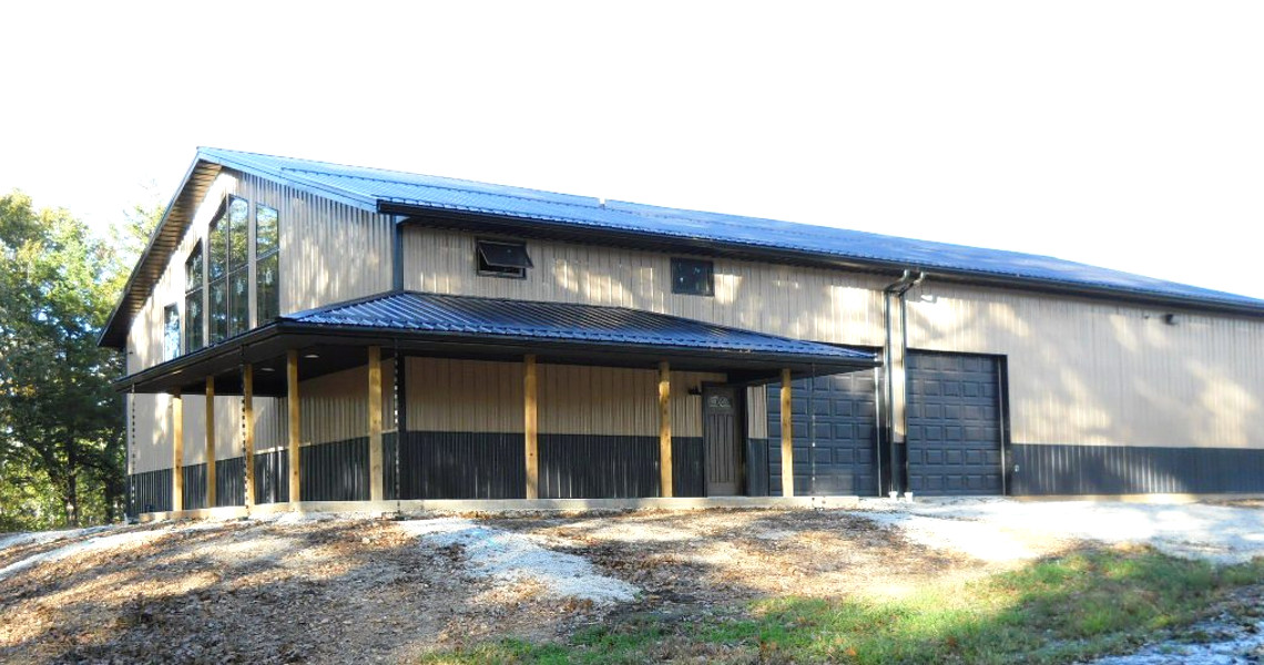 Large metal building with a dark roof, supported by wood columns, perfectly suited for a mountain getaway. It boasts a blend of dark and light siding, multiple large windows on the side, and a wide garage door. Surrounded by a gravel path and trees in the background.