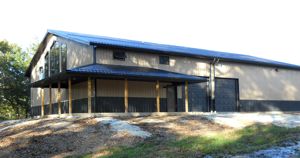 A large, modern barn-style building with a metal roof and wooden paneling serves as a perfect mountain getaway. It features a wide garage door and several windows, surrounded by trees and a dirt driveway. The structure stands on a gentle hillside with sunlight filtering through.