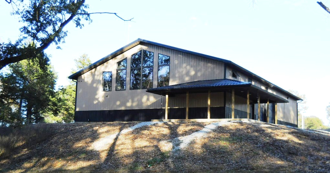 Perched atop a small hill, this modern barn-style building offers a perfect mountain retreat. With large windows and wood paneling beneath a metal roof, it seamlessly blends with the surrounding trees. Sunlight dances on the sloped ground, enhancing the serene nature vacation vibe.