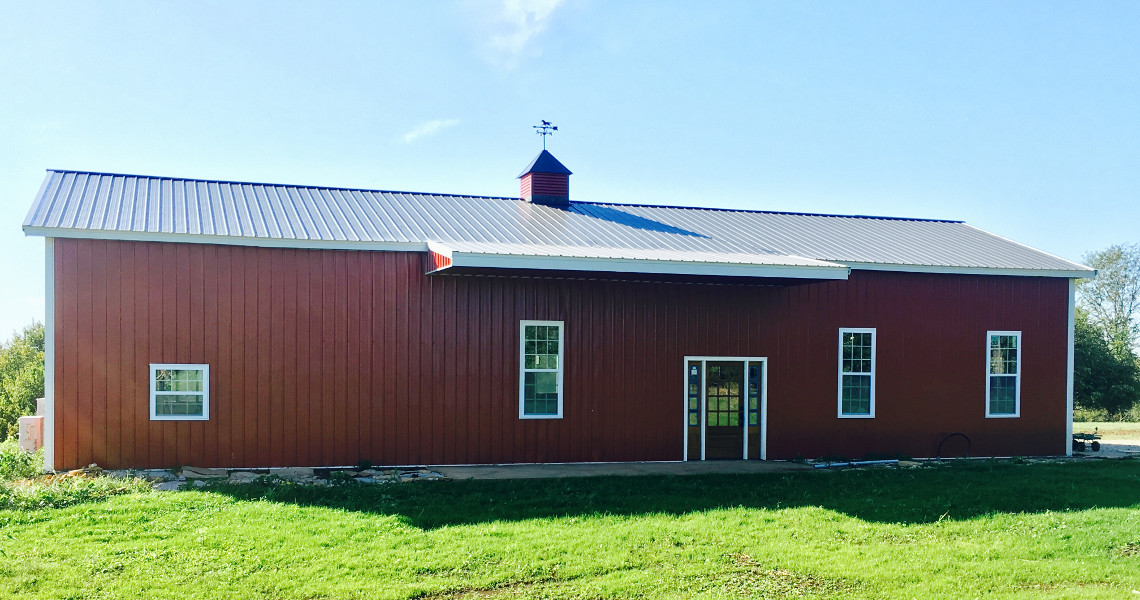 A red barn with a metal roof stands on a lush green lawn under a clear blue sky, resembling a charming barndominium. It features a central entry door, several windows, and a small weather vane on top. Trees are visible in the background.