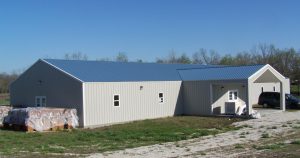 A large metal building with a blue roof, resembling a peaceful church, stands on a grassy area with a gravel driveway. Several crates covered in plastic are near the structure, and a vehicle is parked under a carport. Tall trees frame the scene under the clear blue sky.