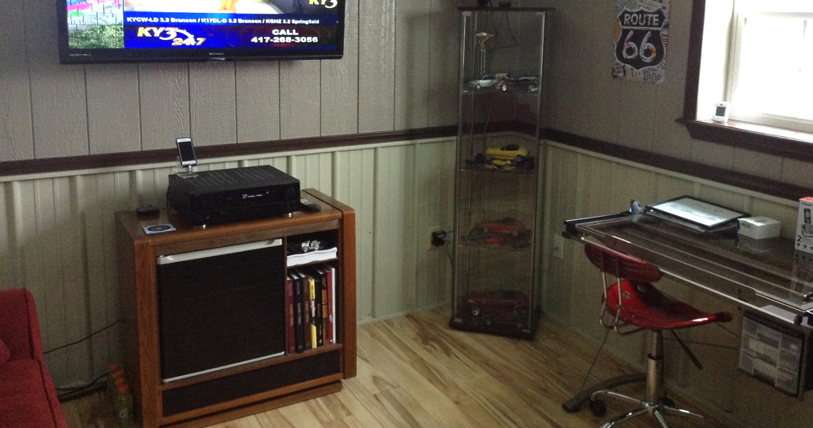 A cozy room corner exudes a country charm, featuring a wall-mounted TV, a cabinet with books and electronics, and a tall glass display case with model cars. A small desk with a red chair sits beneath the warm wood paneling and a nostalgic Route 66 sign.