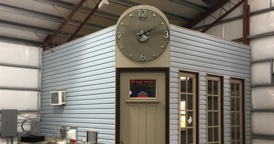 The interior view of the warehouse reveals a small office structure with country charm. The office, adorned with blue siding and a large clock above the door, is lined with windows on one side. A sign graces the door, while an air conditioning unit provides comfort in this industrial paradise.