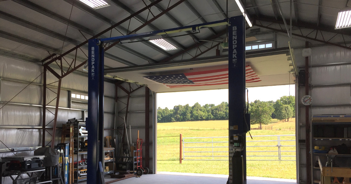 A spacious garage with a blue car lift and an American flag hanging from the open entrance offers a slice of country paradise. The open door reveals a view of green fields and trees under a clear sky, while shelves and tools are visible on the left side.