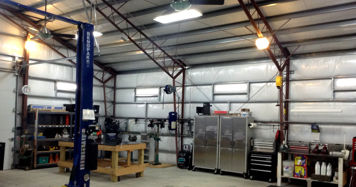 A spacious, organized workshop with metal walls offers a craftsman's paradise, featuring a vehicle lift, workbench, metal cabinets, and various tools. The ceiling boasts exposed beams and overhead lights, ensuring the space is well-lit and tidy.