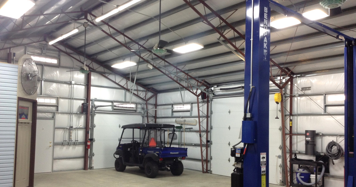 A spacious garage with a blue vehicle parked inside feels like an automotive paradise. The space features a high metal ceiling, large garage door, industrial fan, and blue lift system. Shelving and tools line the walls, creating an organized country haven for car enthusiasts.