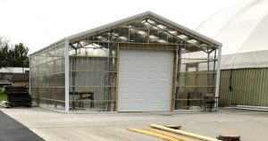 A transparent greenhouse with a white roll-up door, nestled on a paved surface, showcases clear panels and visible wooden beams. Nearby at Bockelman's Garden Center are stacks of wooden planks and a partially visible large dome. Trees and a building frame the picturesque scene.