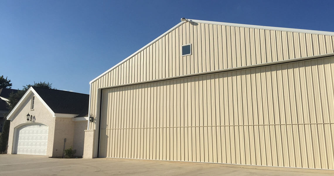 A large beige metal building, complete with steel building accessories and a wide sliding door, stands beside a smaller white house with a three-car garage. The scene is set under a clear blue sky.