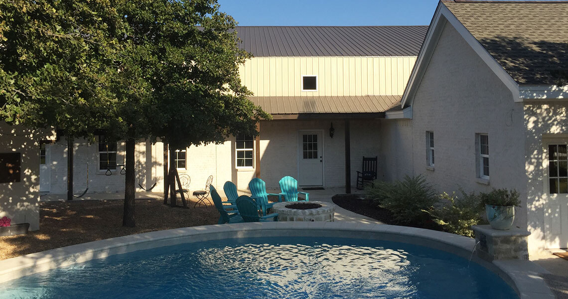 A serene backyard features a round pool in the foreground and a fire pit surrounded by turquoise chairs. A swing hangs from a tree near a white building with a metal roof, under a clear blue sky.