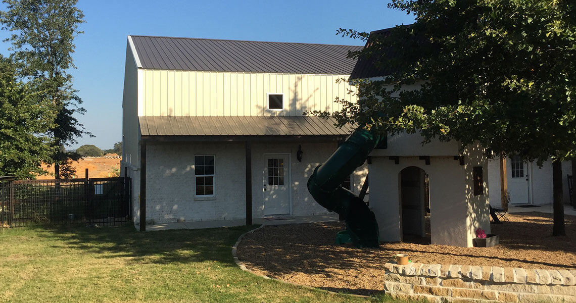 A two-story building with a metal roof and white walls is partially shaded by a tree. A green spiral slide is attached to the structure, descending from the second floor. The area is surrounded by grass and a stone-lined walkway.