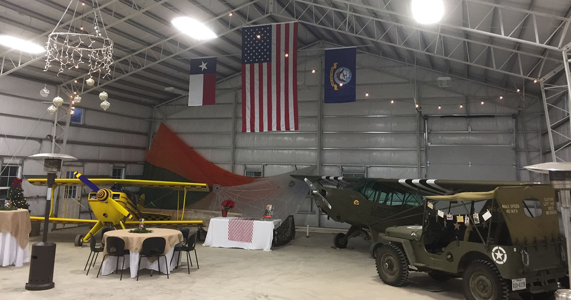 A hangar interior with two vintage airplanes and a military jeep. Tables with chairs are set up for an event. Flags of the United States and other states hang on the wall. Ceiling lights and a small decorated Christmas tree add to the ambiance.