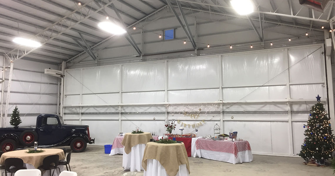 A spacious indoor venue with white walls hosts a gathering. Decorated tables with tablecloths are set up, along with a buffet. A vintage car is parked to the side, and a Christmas tree with lights adds festivity to the scene. Hanging lights illuminate the area.