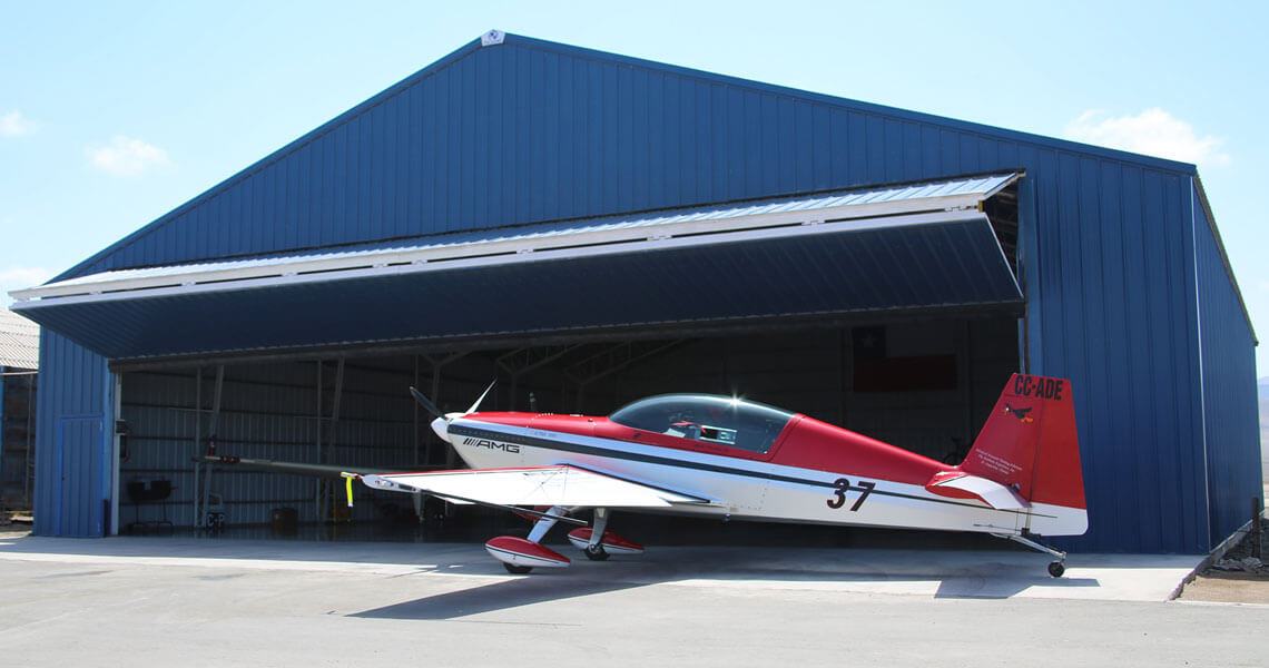 A small red and white aircraft with the number 37 on its side is partially outside a large blue hangar with its doors open. The aircraft is parked on a concrete surface under a clear blue sky.