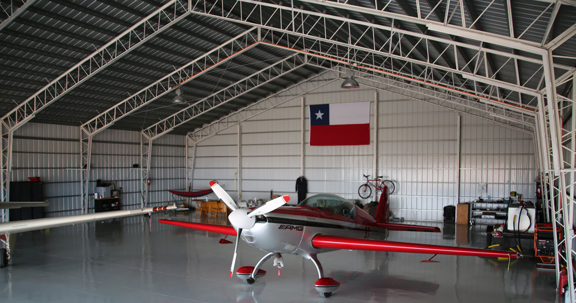 A small red and white aircraft is parked inside a hangar with a shiny floor. The interior features a Texas flag on the back wall, a bicycle, and various equipment and tools along the sides.