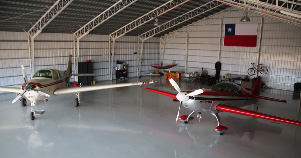 Inside the expansive airplane hangar, two small aircraft rest beneath a metal roof. A Texas flag adorns the back wall, and a red bicycle hangs nearby. Various equipment is scattered throughout the space, adding to the hangar's industrious charm.