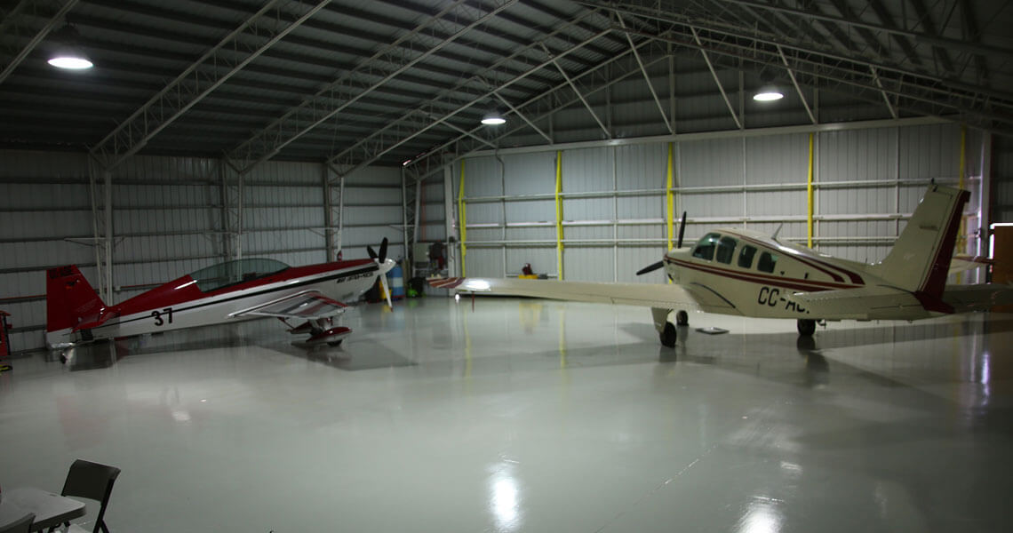 A large indoor hangar with two small airplanes parked inside. One plane is red and gray with the number 