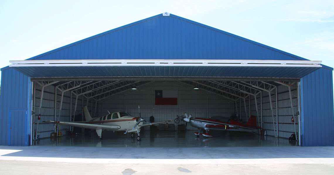 A blue aircraft hangar with its doors open, housing three small airplanes. The hangar has a high, peaked roof and the interior is visible. A flag is displayed inside at the back wall.