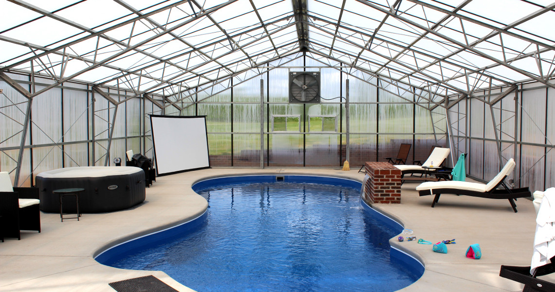 An indoor pool with a small jacuzzi, lounge chairs, and a brick fireplace under a greenhouse-style roof. A projector screen is set up at one end. The area is well-lit with natural light.