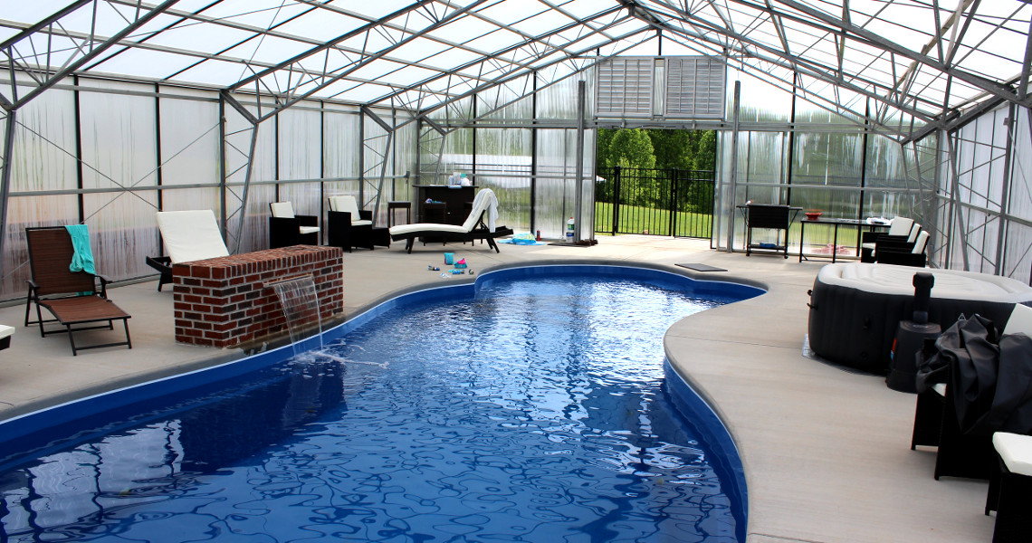The pool house boasts a blue kidney-shaped indoor pool, surrounded by lounge chairs and a hot tub. It features a glass roof and walls for natural light. A charming brick waterfall sits nearby, with towels and toys adding to the inviting ambiance.
