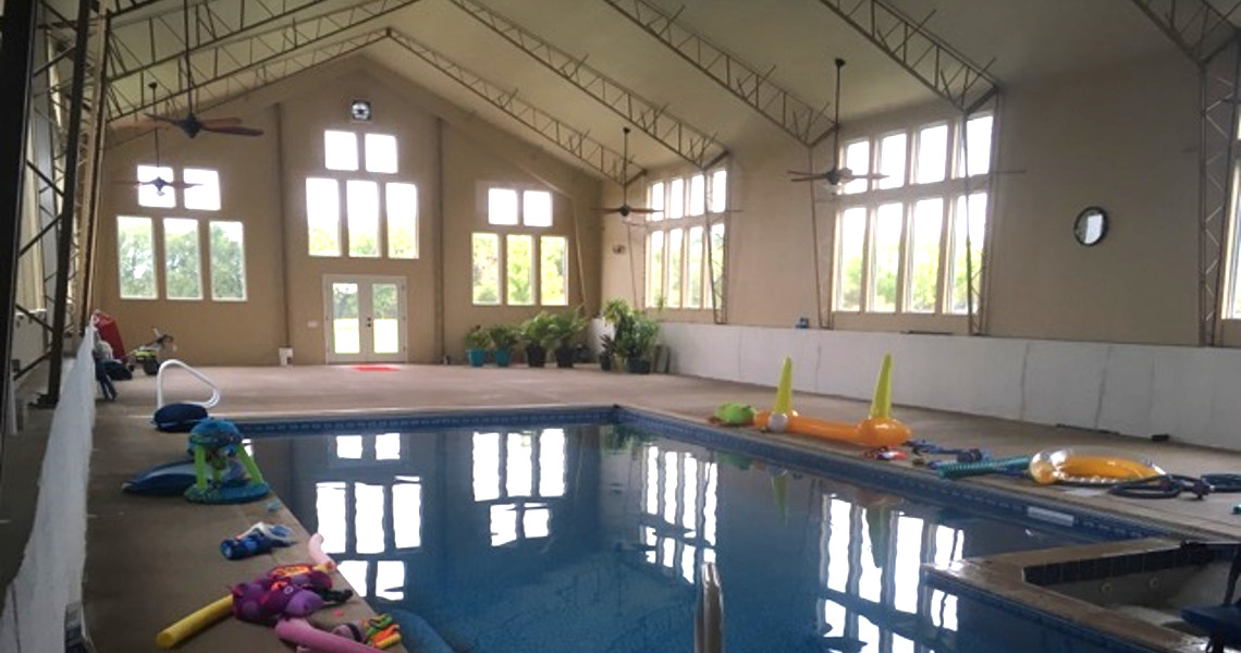 An indoor pool house with inflatable toys scattered around offers a spacious and inviting escape. The big pool is illuminated by natural light streaming through large windows, while potted plants add a touch of greenery near the entrance, creating a pristine and airy atmosphere.