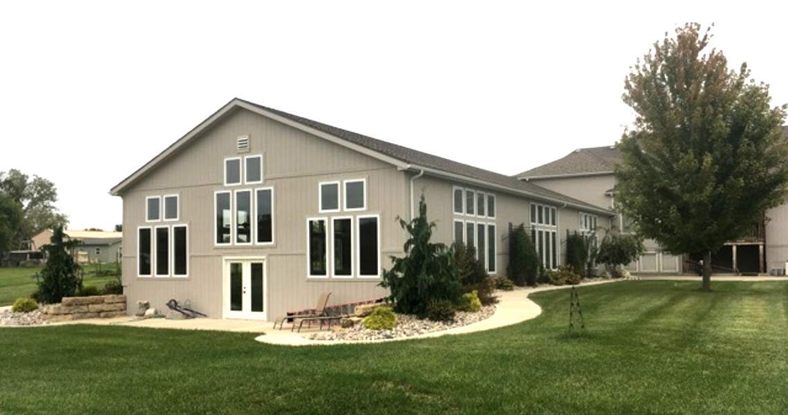 A beige house, known as the Big Pool House, features large windows and is surrounded by a well-maintained lawn and small trees. A curved pathway leads to the entrance. The sky is overcast, with a mature tree standing to the right.