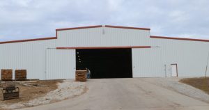 A large white industrial warehouse with an open entrance. Two people stand near the entrance, surrounded by stacks of wooden pallets and various building materials. The sky is overcast, adding a gloomy backdrop to this bustling garrison of activity.