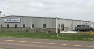 A large, single-story industrial building features a sign that reads "Nemo Mfg, Inc. Electronic Contract Manufacturing" on the front. The gray exterior with brick accents complements a van parked outside, while a grassy area spreads across the foreground.