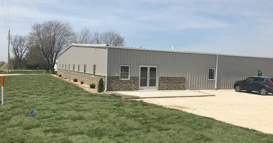 A large, gray metal building with brick accents on the lower walls sits on a grassy area, embodying the essence of industry. Several small bushes line the side, while a few trees linger in the background. A gray car is parked to the right on a gravel area, perhaps hinting at its role in manufacturing for Nemo Mfg.