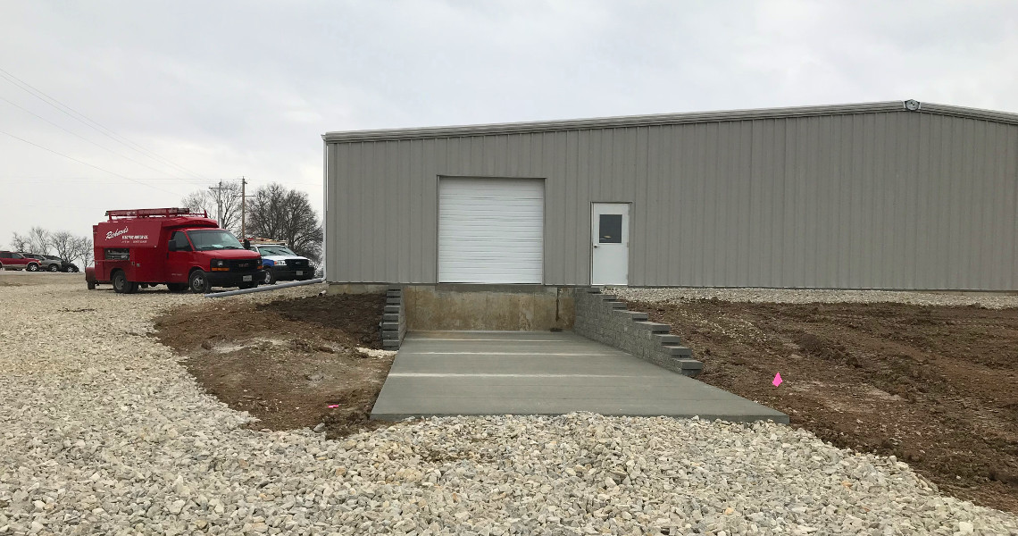 A newly constructed warehouse with a closed white garage door, side door, and a concrete loading dock in front displays the efficiency of Nemo Mfg. Nearby, a red utility truck and other vehicles are parked on a gravel lot under an overcast sky.