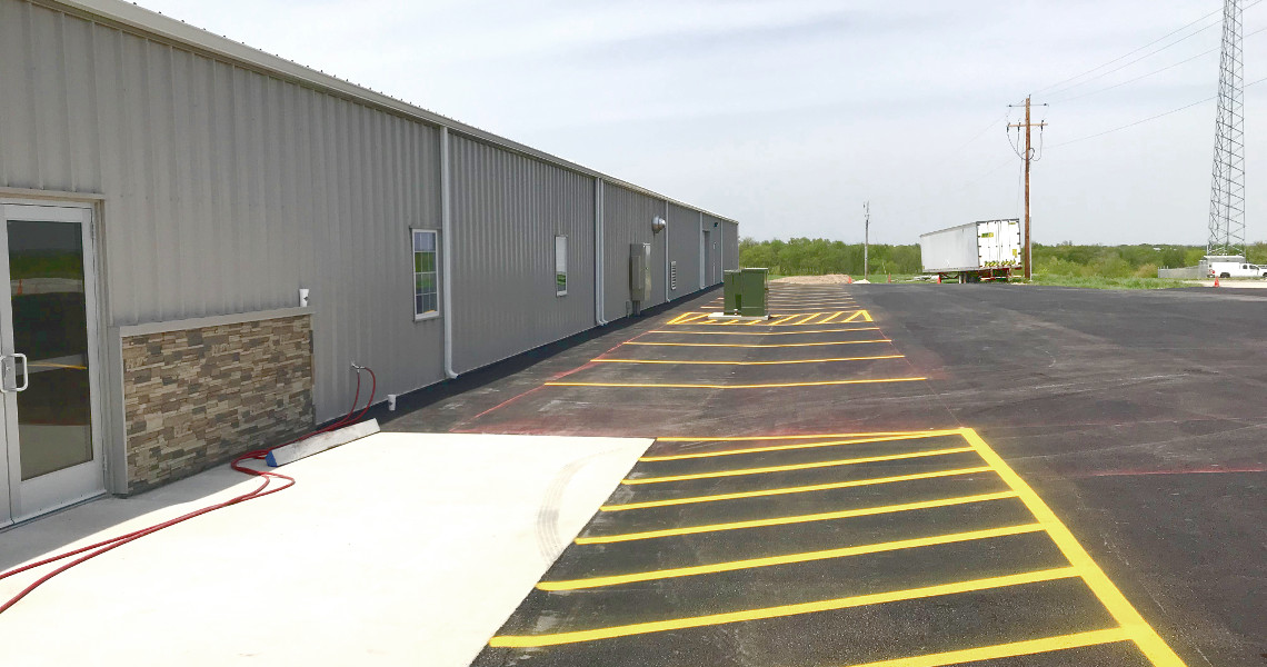 The exterior view of the Nemo Mfg industrial warehouse showcases sleek metal siding, complemented by a paved asphalt parking lot with distinct yellow-striped spaces. A few utility structures and electrical poles stand silhouetted against the clear sky, emphasizing its modern aesthetic.