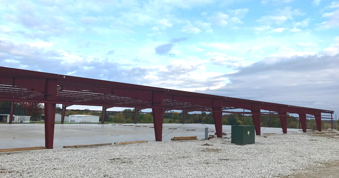 A construction site featuring a large red steel frame by Nemo Mfg rests on a gravel foundation. The sky is partly cloudy, surrounded by trees in the background, with wooden planks and a green utility box adding to the scene's industrial charm.