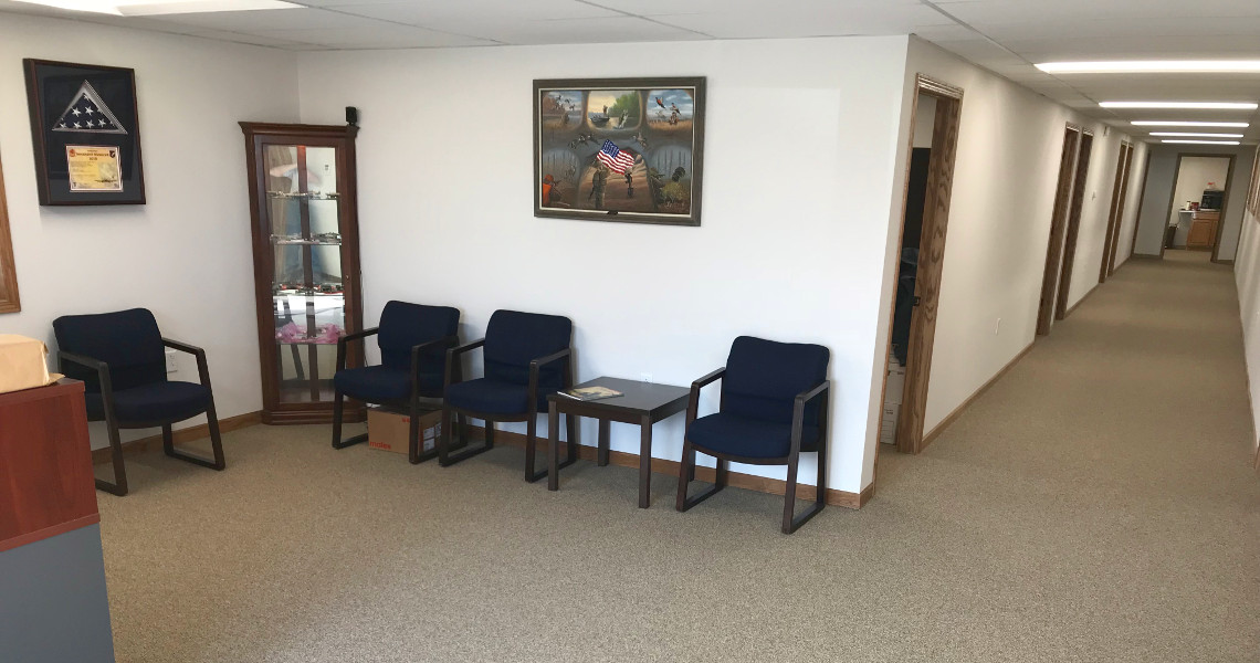 A waiting room with four blue chairs surrounds a small table, accompanied by a display cabinet filled with memorabilia from Nemo Mfg. A framed picture adorns the wall, and a long hallway extends to offices in the background, though it lacks sufficient content for identifying SEO keywords.