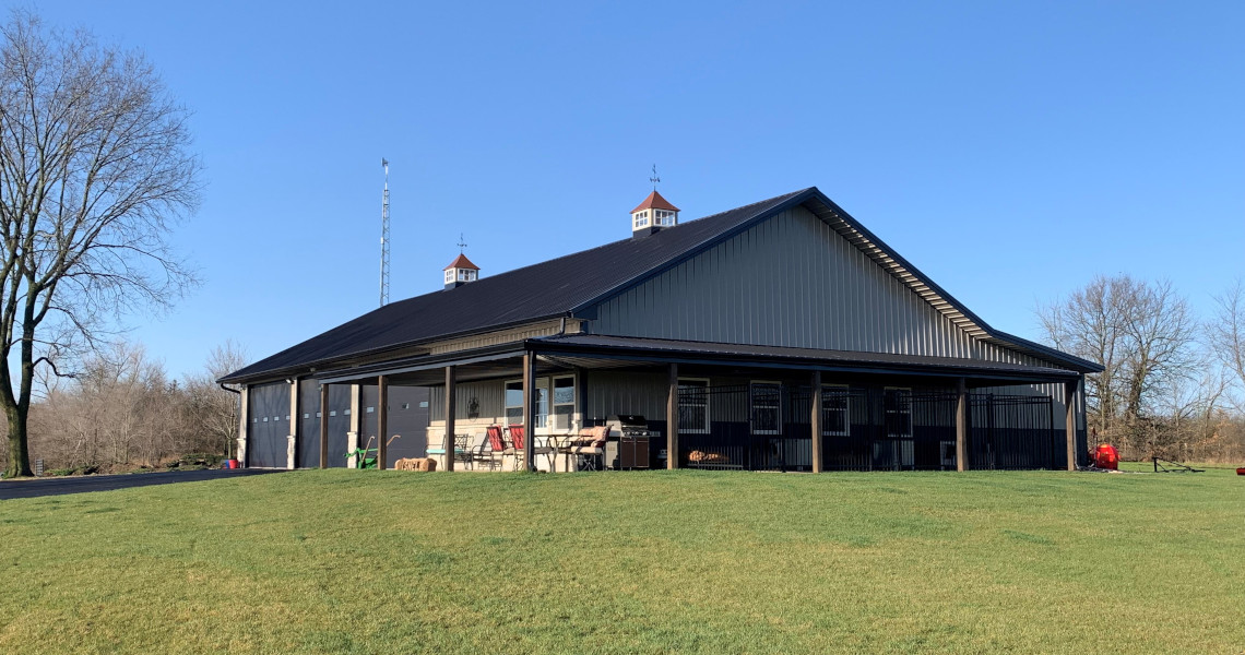 A large, gray barn with a black roof stands in an open field under a clear blue sky. Two small cupolas are on the roof. The area around the barn is grassy with a few bare trees visible in the background.