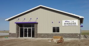 A single-story beige building labeled "Spencer Flight & Education Center" features a stone facade and purple accents around the entrance, embodying the spirit of a premier flight school. A clear blue sky serves as the backdrop.