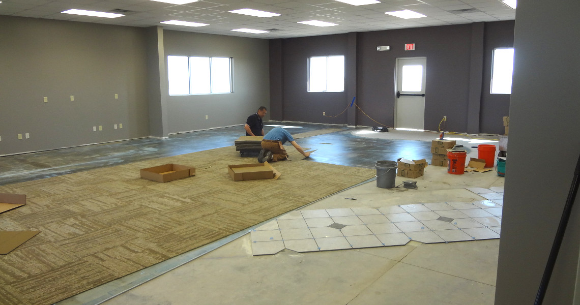 Two workers install carpet tiles on the floor of a large, partially finished room that could easily double as a spacious classroom for aviation training. The area is lit by fluorescent lights and natural light from several windows, with various tools and materials scattered around the room.