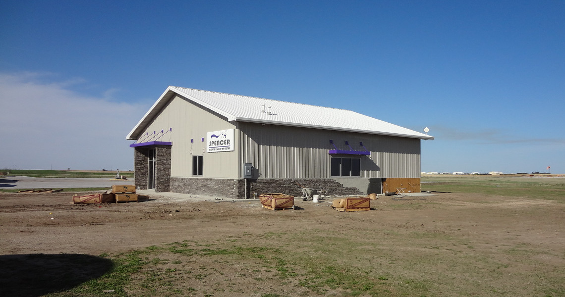A small gray building with a gable roof and purple accents stands on a grassy field, near the bustling Flight School. Labeled 