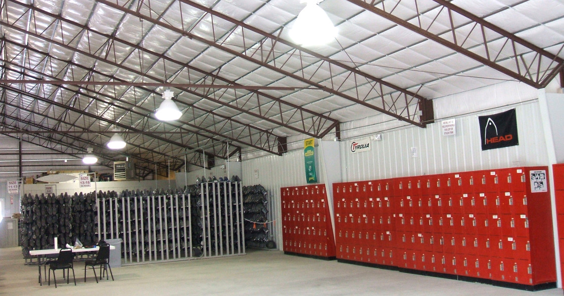 A large indoor space at Snow Creek features red lockers lining one wall, with metal racks filled with skiing equipment on the opposite side. A few tables and chairs sit in the foreground, while exposed metal beams and bright lighting adorn the ceiling.
