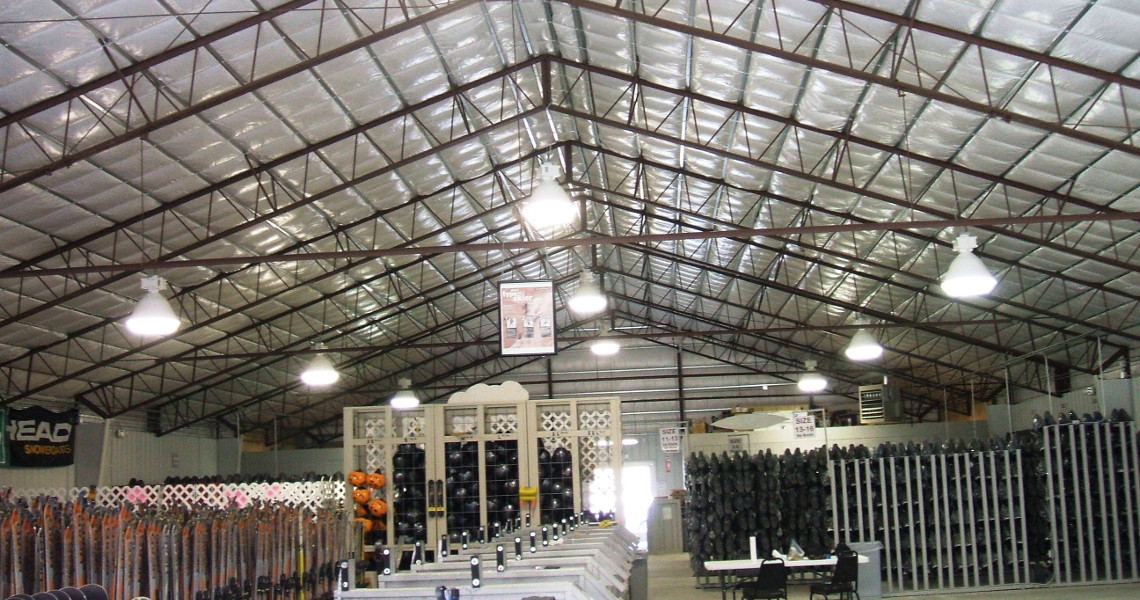 The interior of the Snow Creek warehouse is vast and well-lit, with a metal roof sheltering rows of shelves stocked with equipment and sports gear ready for Ski Resort guests. In the foreground, a few long tables are carefully arranged, adding to the organized charm of the space.
