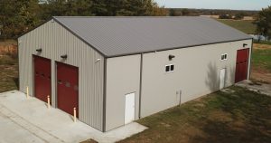 A large, gray metal warehouse resembles a firehouse with two red garage doors and a small white door. The building is surrounded by a grassy area and bordered by a few trees in the background. The sky is overcast.