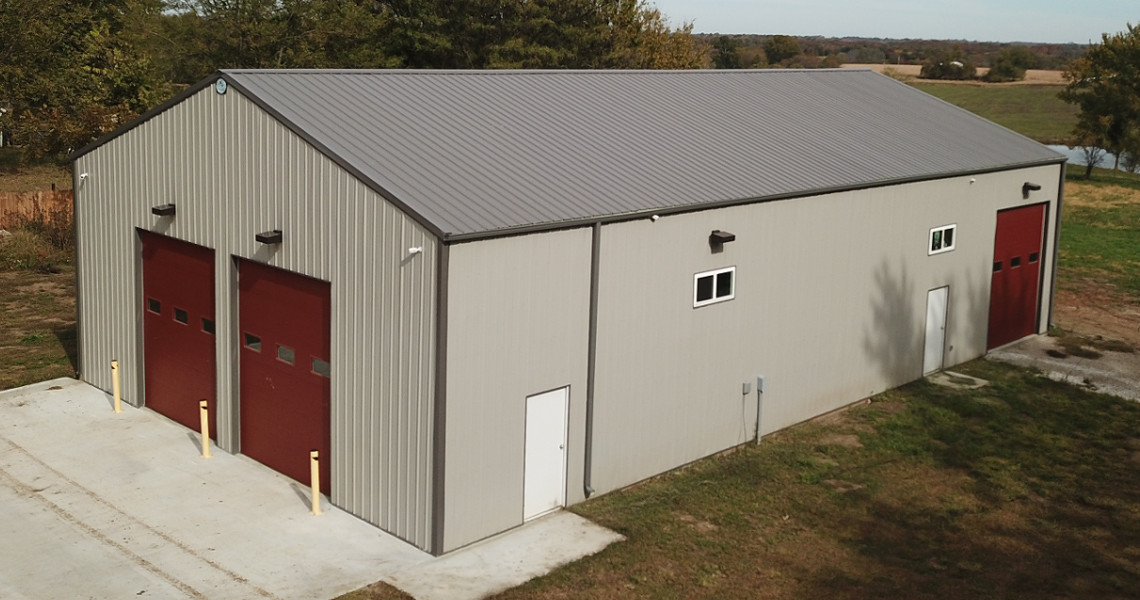 A large, gray metal warehouse resembles a firehouse with two red garage doors and a small white door. The building is surrounded by a grassy area and bordered by a few trees in the background. The sky is overcast.