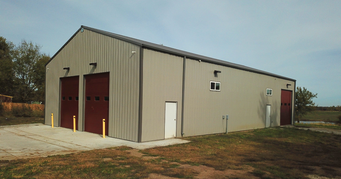 A large gray metal firehouse features two tall red garage doors and a small white door on the side. Situated on a concrete lot with grass surrounding the area, the scene is completed by a clear sky with a few clouds.