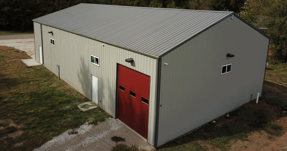 A large gray metal warehouse with a striking red garage door and a few small windows is nestled on a grassy and gravel area. Resembling a firehouse, the building features a sloped roof and fits seamlessly into its rural or industrial setting.