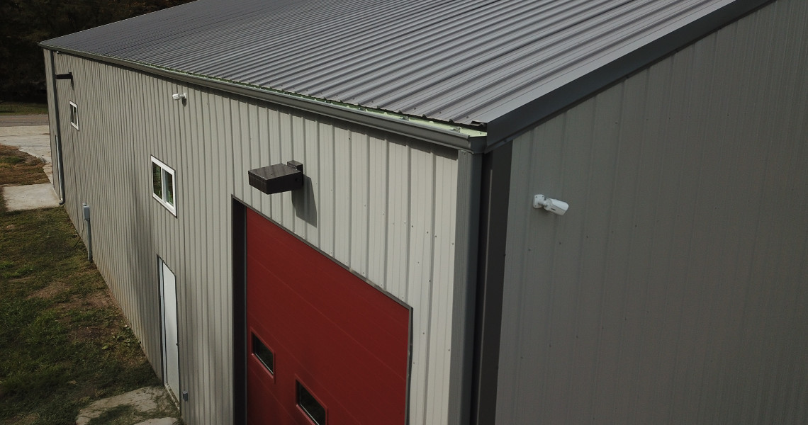 A grey metal firehouse with a sloped roof features a large red garage door with two small windows. A security camera and light fixture are mounted near the door. The building is surrounded by grass and a concrete path.