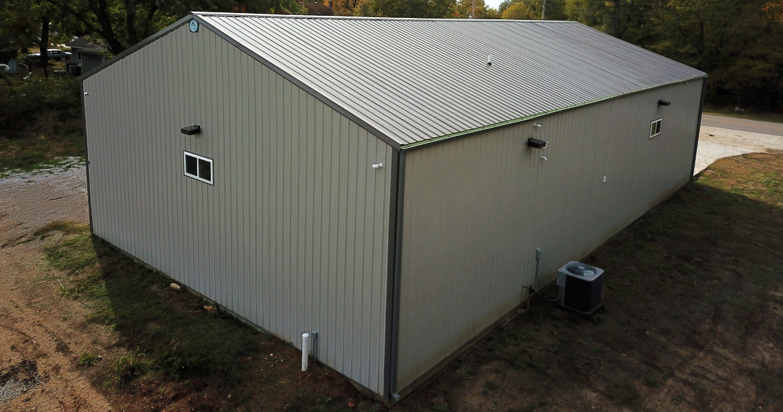 A large, gray metal building resembling a firehouse garage features a sloped roof and is viewed from an elevated angle. The structure, with small windows and doors, sits amid grass and dirt. An air conditioning unit is strategically placed near one of its sides.