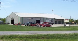 Exterior view of a large gray warehouse, possibly serving as a rental hub, with multiple garage doors and signs. Several cars and equipment are parked in front. A sign on the lawn reads "Rex's Store." The sky is clear, with grass and a road in the foreground.